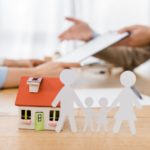White paper cut family and house model on wooden table with blurred people at background, life and